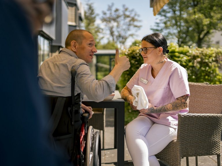 Eine Mitarbeiterin hört einem Bewohner zu, der etwas erzählt. Sie sitzen auf einer sonnigen Terasse.