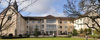 Junge Pflege Rheine Über eine Rasenfläche blickt man auf ein U-Förmiges Gebäude in Beige. Auf dem Dach des linken U-Schenkels befindet sich ein Türmchen mit Kreuz. Mittig angeordnet ist der Eingangsbereich über dem der Schriftzug "St. Josefshaus" zu lesen ist.